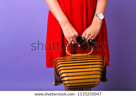 Fashion close up picture or woman wearing elegant bright red dress, holding straw wooden trendy bag and heart sunglasses, simple watch, purple background.