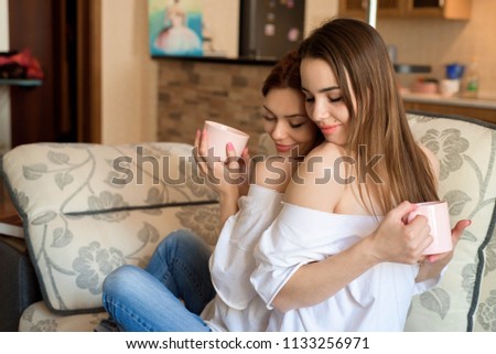 Two sisters on the sofa with a cups of tea in hands. Two best friends enjoying time.