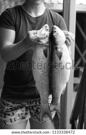 Successful female angler holding two big fish in hands, smiling at camera while having good mood after huge catch at river, posing against blue blank wall. Pretty fisherwoman with cheerful expression