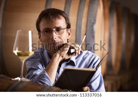 Smiling winemaker in cellar looking satisfied at a glass of white wine during wine tasting, with notebook and pen in hands. Royalty-Free Stock Photo #113337016