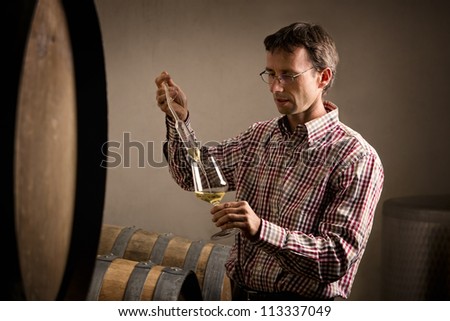 Winemaker in cellar taking sample of white wine from barrel for wine tasting.