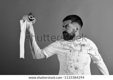 Man with concentrated face on red background. Guy with beard holds jack o lantern and looks at it. Halloween character in white long sleeved ghost costume. October time and spooky holiday concept