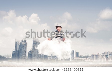 Young little boy keeping eyes closed and looking concentrated while meditating on cloud above wooden floor with cityscape view on background.