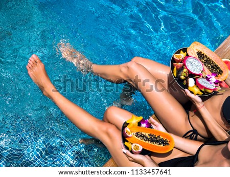 Lifestyle hot close up picture of two woman sitting near pool holding big plates with amazing sweet tasty tropical exotic fruits, amazing long legs, healthy Egan vegetarian lifestyle, diet concept.