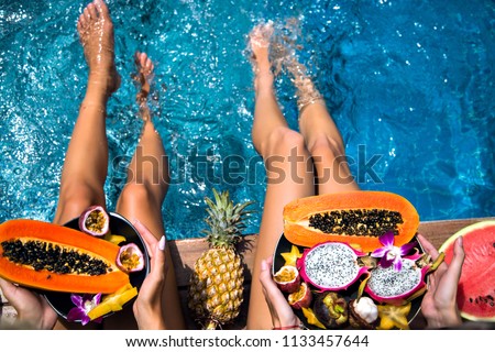 Summertime close up picture of two woman sitting near pool holding big plates with amazing sweet tasty tropical exotic fruits, papaya, mangosteen, dragon fruit watermelon and pineapple, healthy vegan.