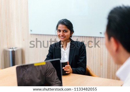An Indian Asian woman has a business meeting (or interview) with a Chinese man. She is speaking animatedly and gesturing with her hands as she talks. She is young, attractive and is wearing a suit.