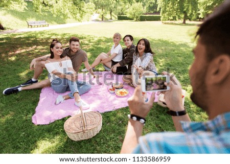 friendship, leisure and technology concept - man taking picture of his friends by smartphone on picnic at summer park