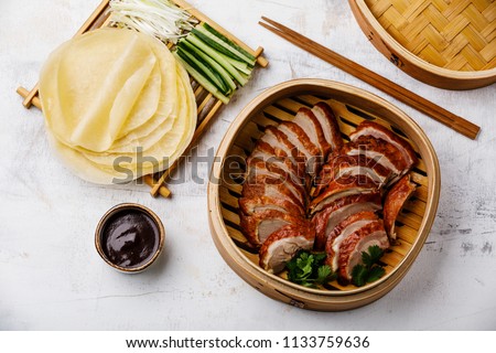 Sliced Peking Duck in bamboo steamer served with fresh cucumber, onion, Hoysin sauce and roasted wheaten pancakes on white wooden background