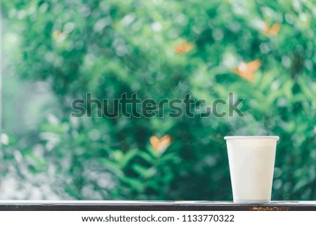 A selective focus picture of a paper cup with hot coffee on wooden table in green leaf garden and smoke and heart bokeh background