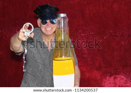 PHOTO BOOTH. A man wears a Police Hat, Holds Hand Cuffs, A Giant Beer Bottle and smiles for his picture in a Photo Booth with a red velvet background.
