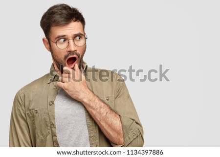 Puzzled anxious bearded male holds chin and looks with frightened expression aside, being terrified by high prices, opens mouth widely, wants to buy new outfit. Blank space for advertisement