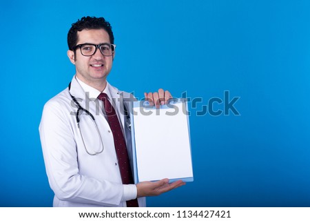 Standing young doctor holding a clipboard showing it smiling, on a blue background