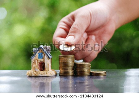 close up hand holding coin, stack of money and toy house on table, green nature background, saving money for building, manage for success business concept