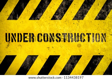 Under construction warning sign text with yellow black stripes painted over concrete wall coarse facade as texture background. Concept for do not enter the area, caution, danger, construction site.