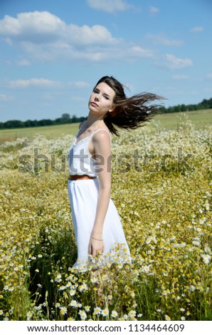 Young female model in outdoor photo session in Poland. Girl in long white dress stands in meadow surrounded by colorful flowers.