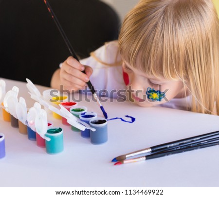 Beautiful little blonde girl, has serious sad face, pretty eyes, white t-shirt. Painted brushes water colors in skin. Child portrait. Kids concept hobby.