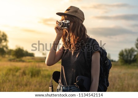 Young female tourist taking photo at hiking trip. Woman with backpack stands at sunset and photographs beautiful rural area