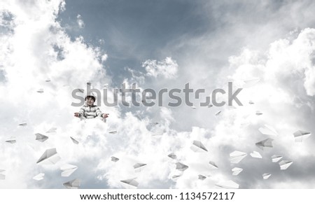 Young little boy keeping eyes closed and looking concentrated while meditating among flying paper planes in the air with cloudy skyscape on background.