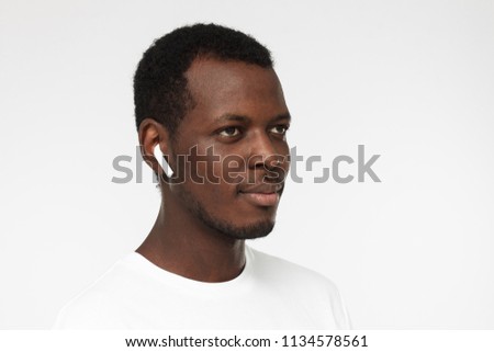 Horizontal headshot of young African American man pictured isolated on grey background with short hair and white wireless earphones listening to audio tracks or conversation with concentrated face
