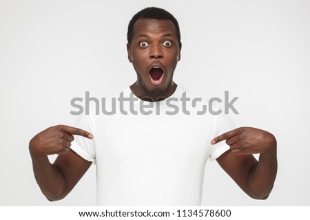 Closeup picture of emotional African American man isolated on gray background drawing attention to content on copy space of blank T-shirt he is wearing with shocked face, advertising something great