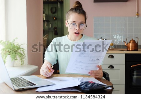 Horizontal closeup of European Caucasian lady spending afternoon at kitchen table, wearing eyeglasses, looking shocked with what she sees in documents she is dealing with, dissatisfied with debts Royalty-Free Stock Photo #1134584324