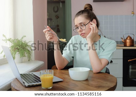 Horizontal portrait of attractive Caucasian girl sitting in kitchen in morning looking tired while eating breakfast, her laptop lying open on table, not sure what to do during day, feeling bored