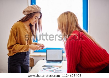 Positive female blogger making photo of desktop via camera on modern smartphone device connected to internet, two multicultural successful designers spending time together for course work indoors