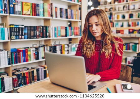 Charming female student making research for startup project using internet information from laptop device in university library, hipster girl typing email for asking job vacancy via app on netbook