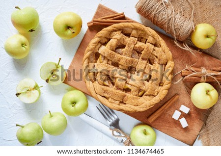 Homemade healthy american apple pie on the brown wooden board with fresh apples, cinnamon, sugar cubes, knife and fork on the white background. Top view