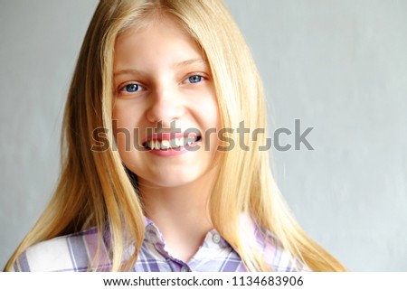 Headshot of beautiful teenage girl with blonde straight hair, blue eyes smiling at the camera. Pretty young female looking amused, wearing white purple plaid shirt. Background, copy space, close up.