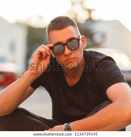Stylish beautiful young hipster man with a hairstyle in sunglasses and a black T-shirt at sunset sits on the street