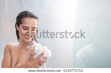 Portrait of beaming woman rubbing body with foam while standing under steam of water. Copy space Royalty-Free Stock Photo #1134771752