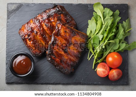 Closeup of pork ribs grilled with BBQ sauce and arugula. Tasty snack to beer on a stone Board for filing on old concrete background. Top view