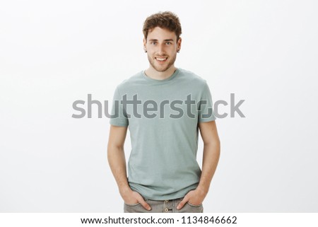 Portrait of charming flirty aduly guy with blond hair, holding hands on pants and smiling broadly, talking with friends casually while hanging out and relaxing, standing joyful over gray wall
