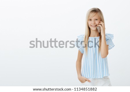 Mom gave daughter cellphone to talk with granny. Studio portrait of positive pleased european child with fair hair in blue blouse, standing casually over gray wall and communicating via smartphone