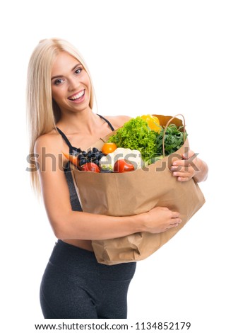 Portrait of happy smiling young beautiful woman in fitness wear, holding grocery shopping bag with healthy vegetarian food, isolated over white background
