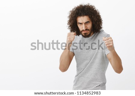 Indoor shot of outraged bully with beard and afro haircut, standing in boxing pose with raised clenched fists, frowning at enemy, being ready to fight, standing angry over gray background Royalty-Free Stock Photo #1134852983