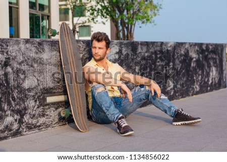 Young handsome man with beard sitting with longboard on the street.
