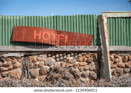 Old and rusted disused hotel sign leaning against a green corrugated fence on top of a stone wall