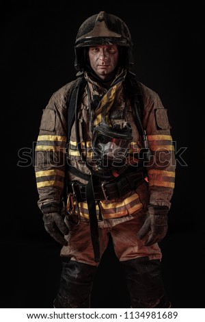 Serious dirty man in uniform of firefighter and helmet standing on black background looking seriously at camera. 