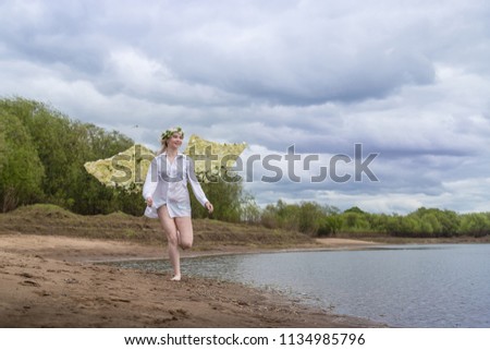 Nice blonde girl with white flower wings on the beach near river in cloudy day