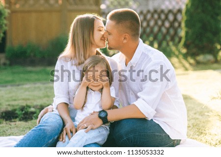 parents kissing while little daughter covering eyes while resting together on backyard
