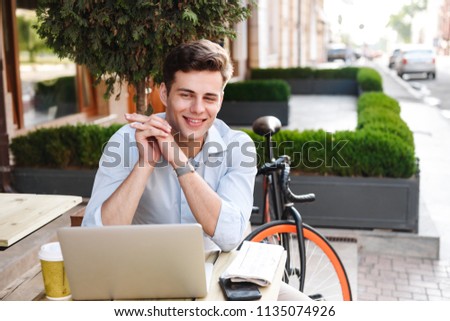 Satisfied young stylish man in shirt working on laptop computer while sitting at a cafe outdoors with bicycle