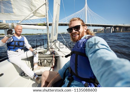 Cheerful handsome friends in blue life jackets taking selfie against beautiful bridge while sitting on boat deck Royalty-Free Stock Photo #1135088600