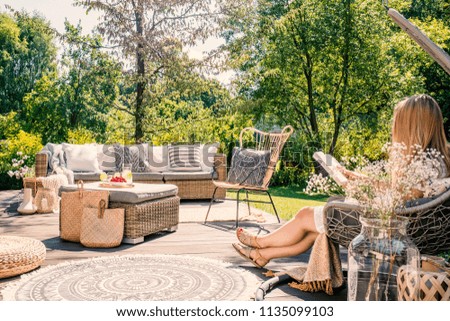 Woman reading book while relaxing at terrace with rattan furniture in the garden. Real photo