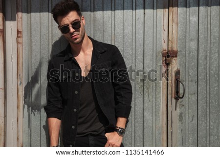 handsome man wearing black clothes and sunglasses standing near garage door with hand in pocket on a sunny day, portrait picture