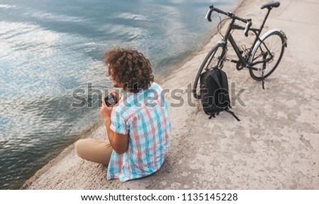 Rear view of young male with curly hair taking photos of nature on his digital camera. Handsome man wears casual shirt relaxing and sitting near to the bike next to the lake. People, travel, lifestyle