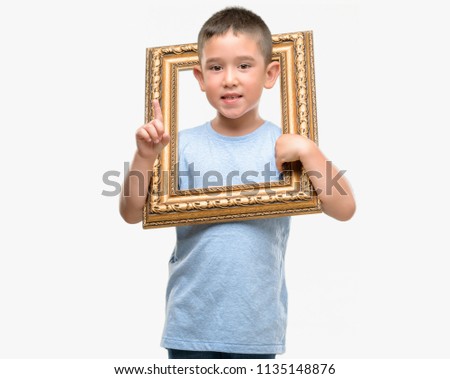 Dark haired little child holding frame surprised with an idea or question pointing finger with happy face, number one