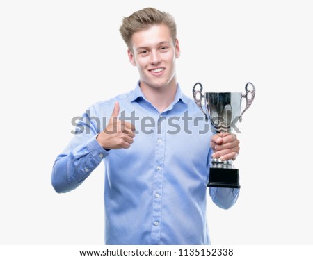Young handsome blond man holding a trophy happy with big smile doing ok sign, thumb up with fingers, excellent sign