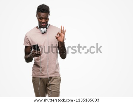 Young african american man wearing headphones and holding smartphone doing ok sign with fingers, excellent symbol